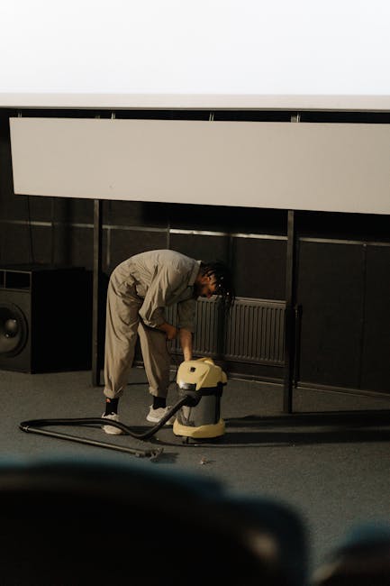 A professional cleaner dressed in full protective white coveralls, gloves, mask, and shoe covers, operating a steam cleaning machine on a beige, textured carpet inside a well-lit, modern living room.